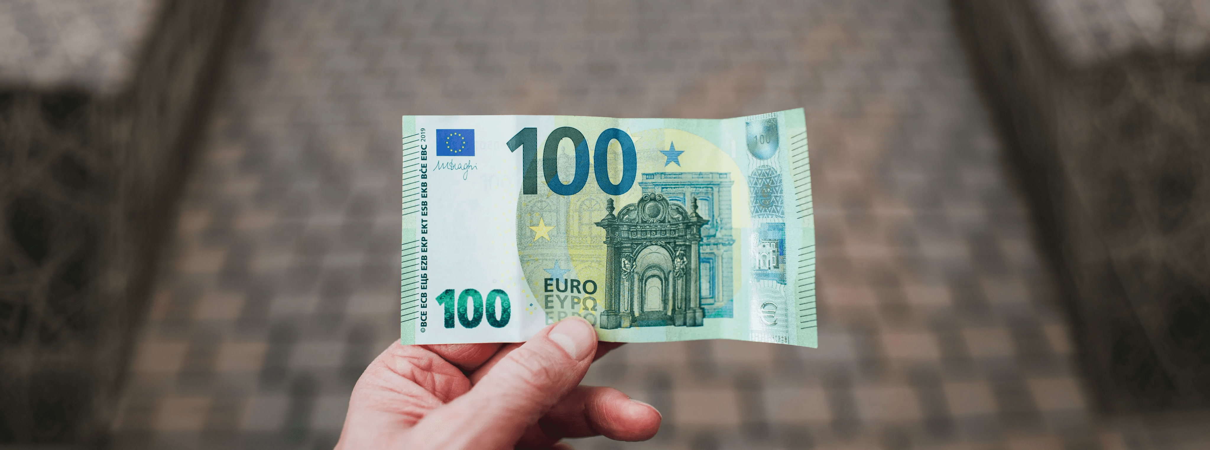 Eine Hand hält einen 100-Euro-Schein in die Kamera vor unscharfem Hintergrund.