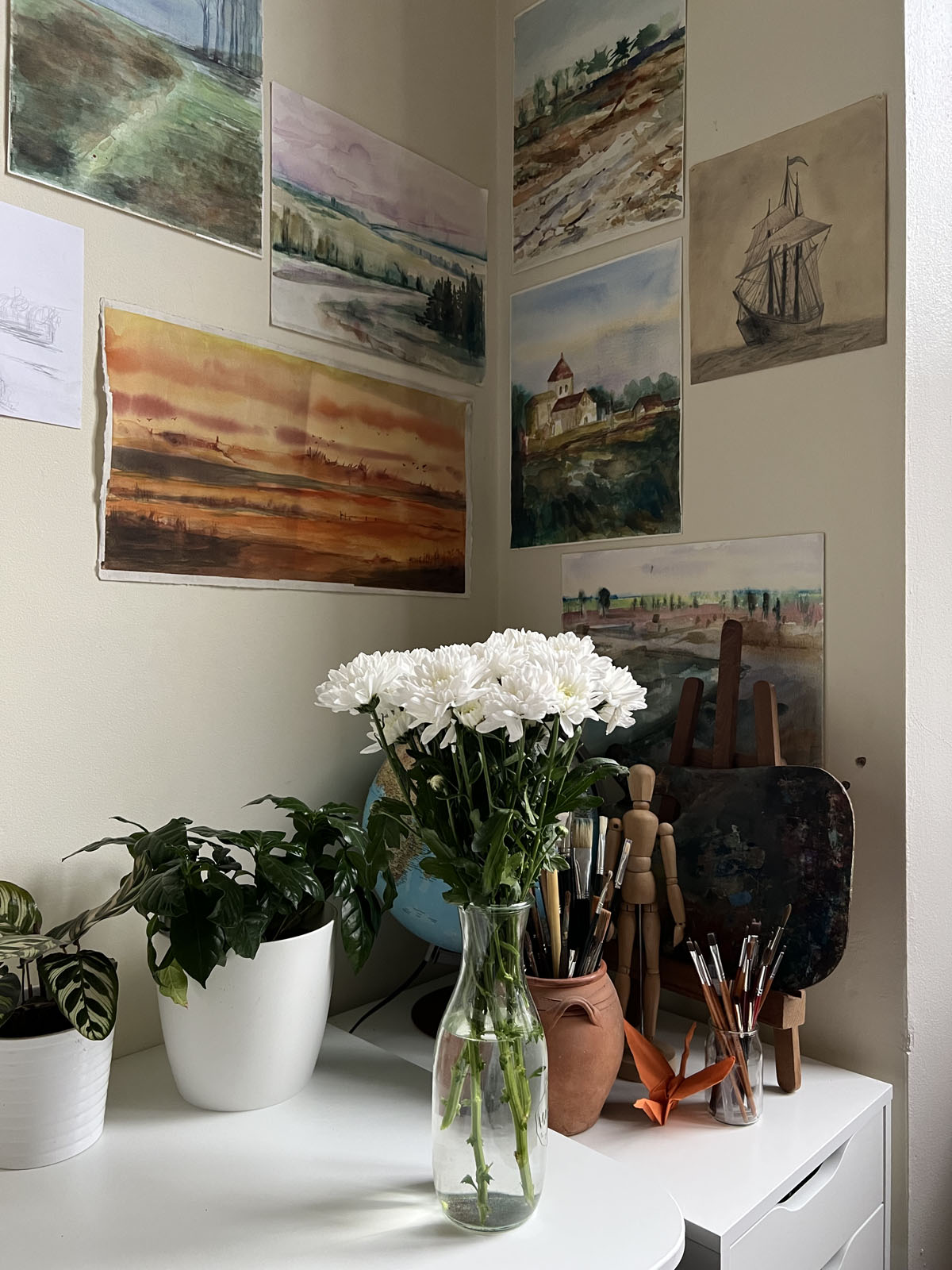 Een sfeervolle hoek van een kunstatelier. Aan de muur hangen verschillende aquarellen, waaronder een groot horizontaal landschap in warme oranje- en bruintinten en een schilderij van een kerkje. Op de voorgrond staat een witte vaas met witte chrysanten, geflankeerd door een groene kamerplant, een houten tekenpop, penselen en een schilderspalet.