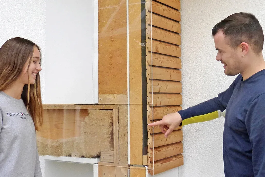Mann zeigt auf eine Schnittdarstellung einer Wandkonstruktion mit Dämmung und Holzverkleidung, eine junge Frau schaut zu.