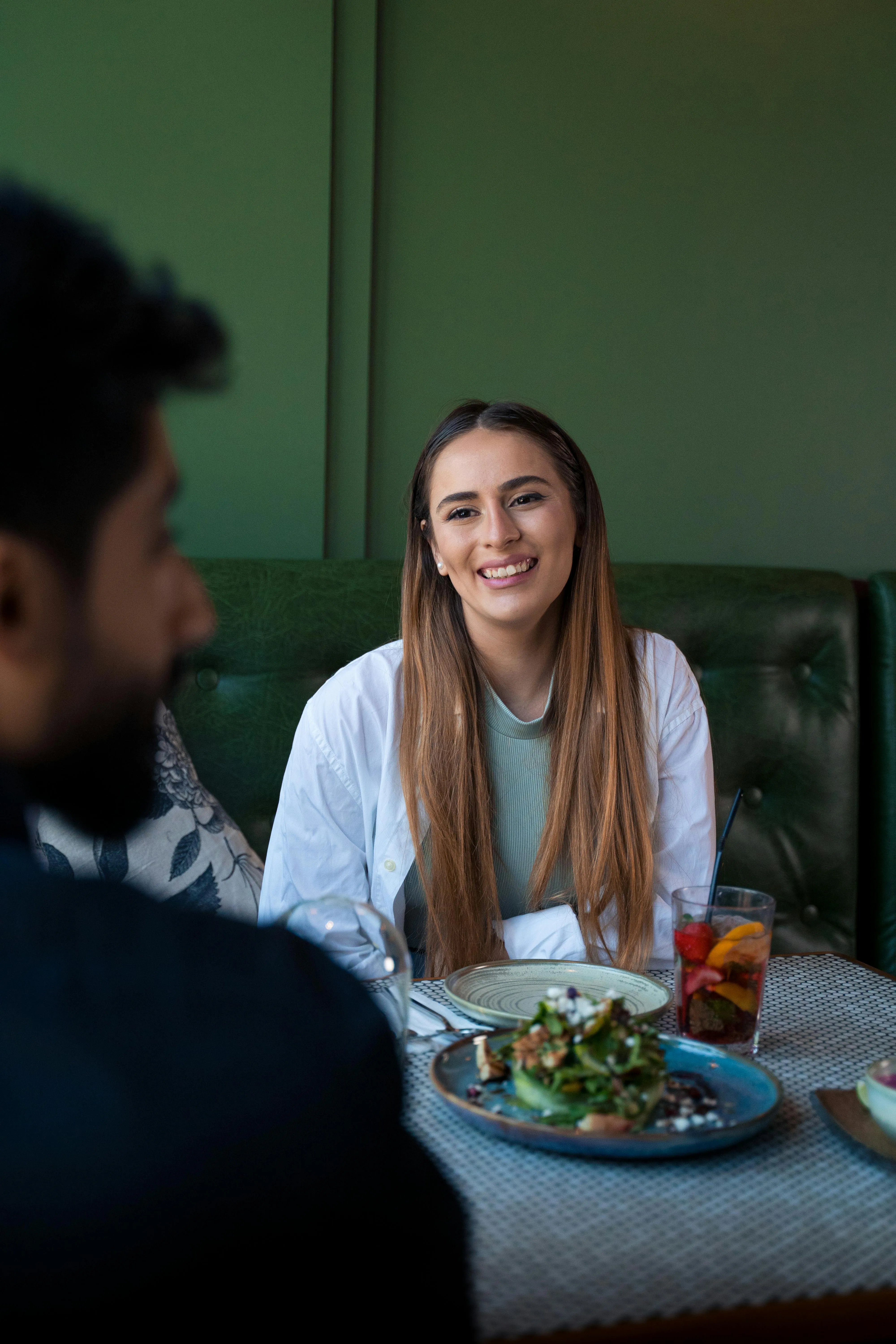 Une femme souriante aux cheveux longs et clairs, portant une chemise blanche ouverte sur un haut vert, est assise à la table d'un restaurant au mur vert. Elle regarde un homme de profil, au premier plan à gauche. Sur la table, on aperçoit une salade et une boisson fruitée.