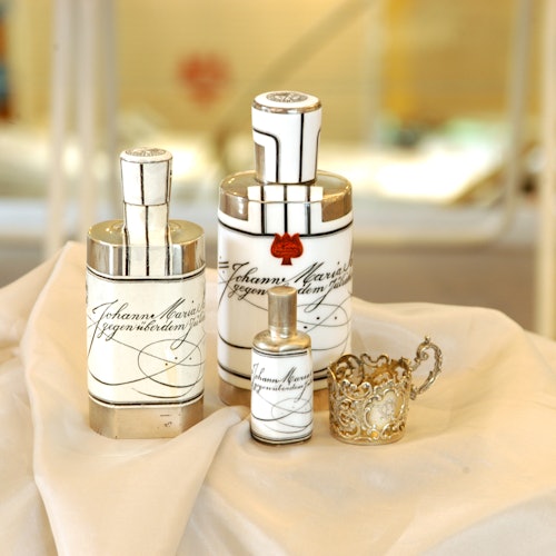 Three elegant white perfume bottles with silver caps and decorative script, arranged on a cream-colored fabric next to a detailed metal holder.