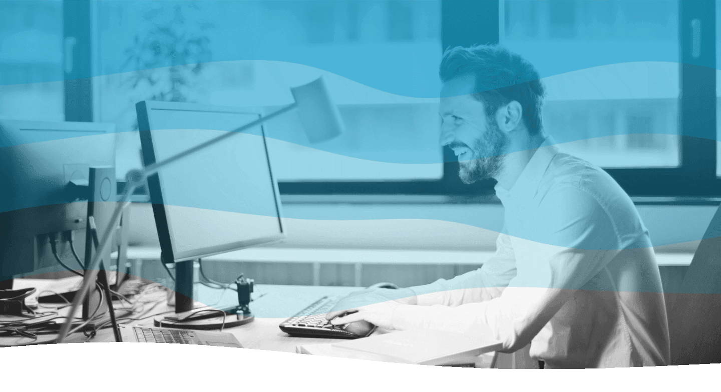 A man with a beard smiles while working at his desk in a modern office, with a large window providing natural light.