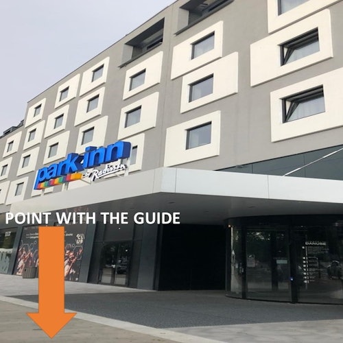 La entrada de un hotel moderno con un letrero de "Park Inn by Radisson", que presenta una flecha naranja etiquetada como "PUNTO CON EL GUÍA."