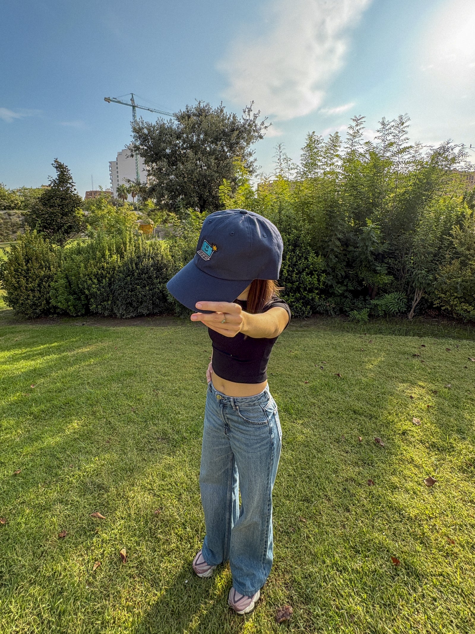 Person wearing a blue cap and jeans standing in a grassy area with trees in the background