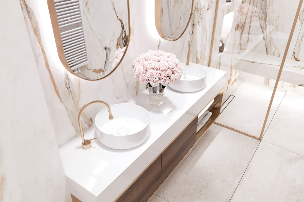 Modern bathroom with marble walls and countertops, two oval mirrors, dual white vessel sinks with gold faucets, and a vase of pink roses centered between the sinks