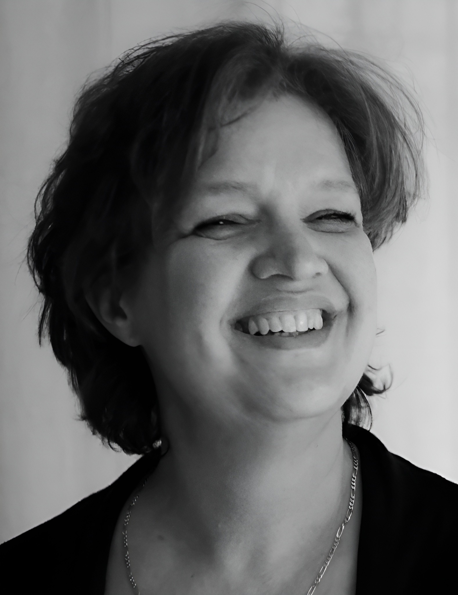 A young person with curly hair smiles at the camera while sitting in front of a laptop against a dark background.