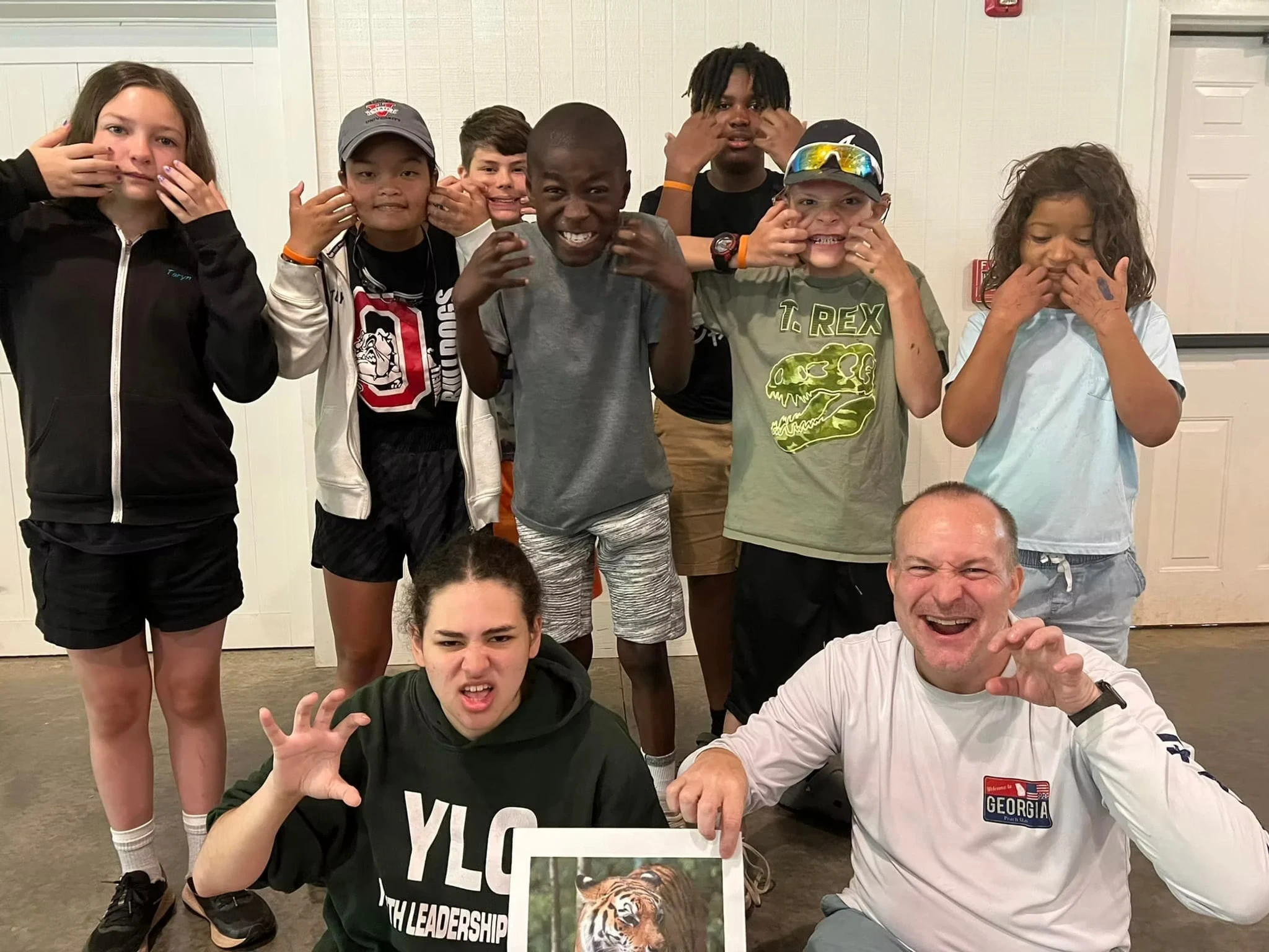 A group of campers and staff playfully signing TIGER, with one adult holding a picture of a tiger.