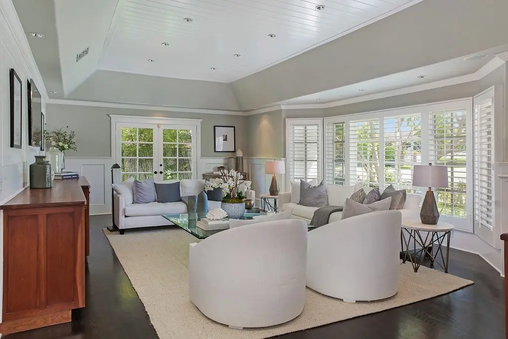 Elegantly remodeled living room with vaulted ceilings, white sofas, gray accents, and large windows with shutters, offering a bright and inviting atmosphere.