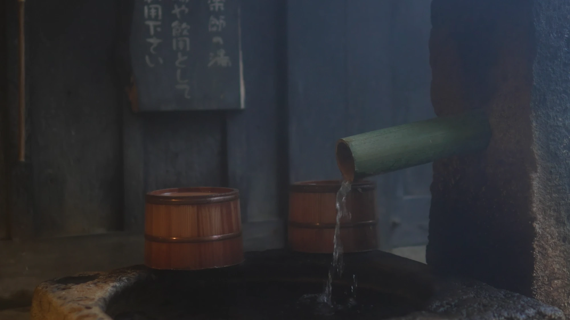 A bamboo spout running into a dark bathing tub at Kurokawa Onsen, wooden buckets beside it and a posted notice on the wall