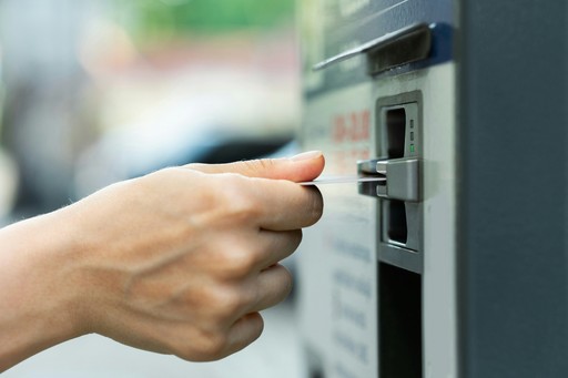 A close-up side view of a person's hand inserting a white card into the slot of a gray electronic payment machine or parking meter.