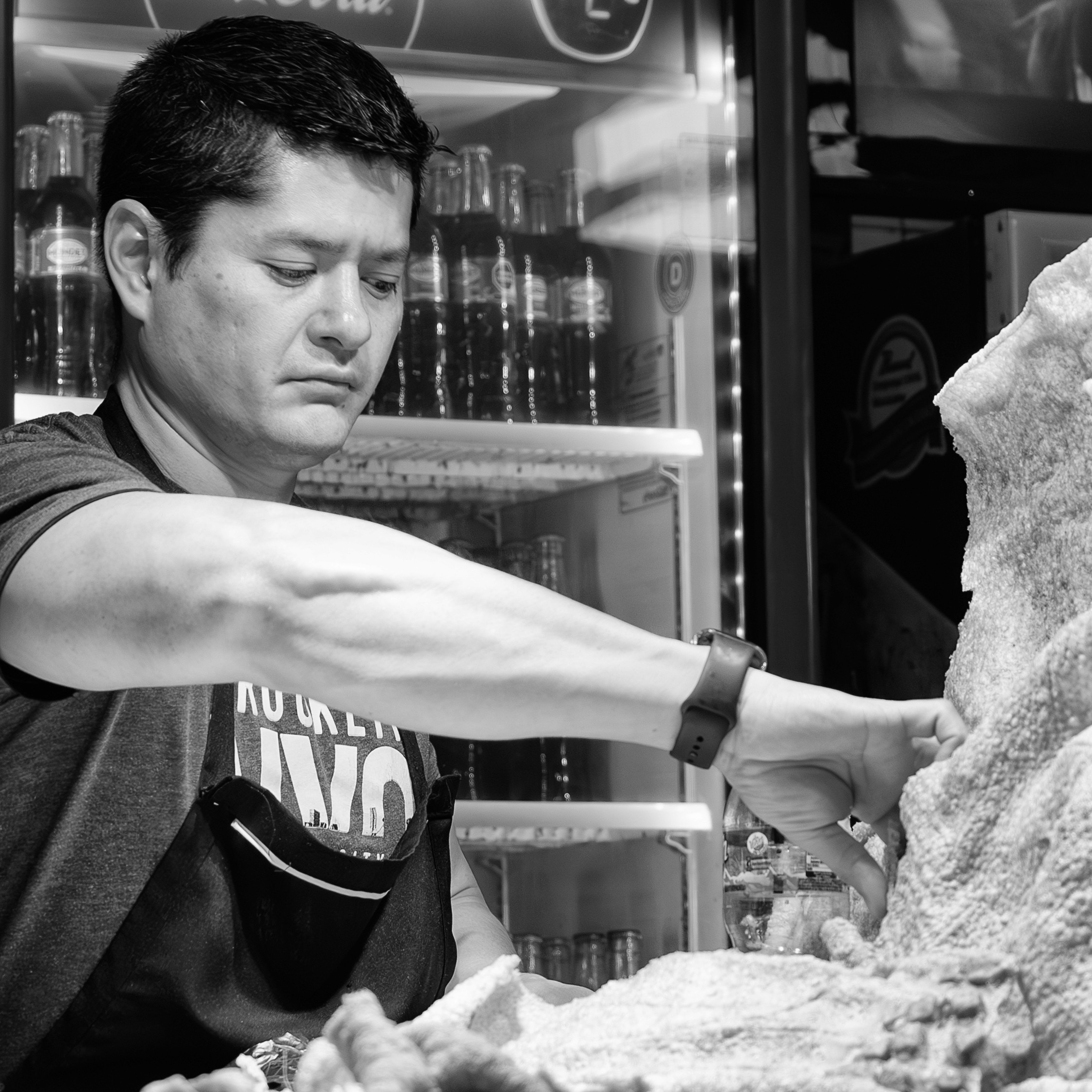 Black and white photo of a vendor carefully arranging large pieces of chicharrón in a market setting, with a fridge of glass soda bottles in the background.
