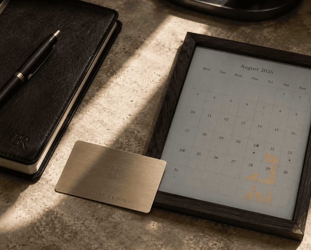 A top-down desktop arrangement bathed in warm natural sunlight streaming across a textured beige stone counter. A framed digital E-Ink calendar display in a dark wooden frame shows an August 2026 grid layout. Resting beside it is a brushed gold metallic card engraved with the "BR" monogram and "Blackthorne & Rowe." On the left sits a dark brown leather-bound journal embossed with the same monogram, paired with a sleek black and gold executive pen.