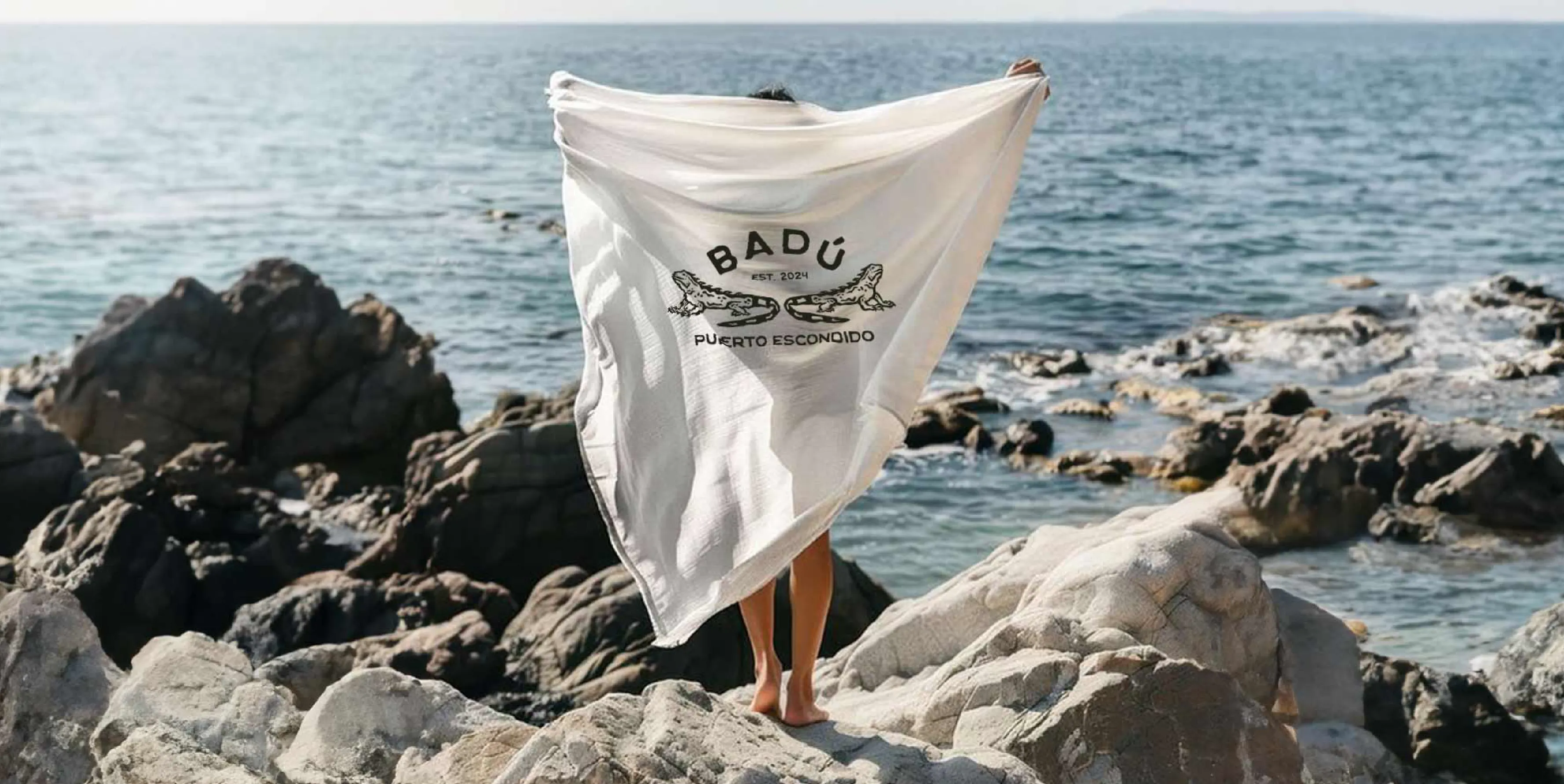 Badu printed logo on a beach towel, held up by someone right on the edge of the beach