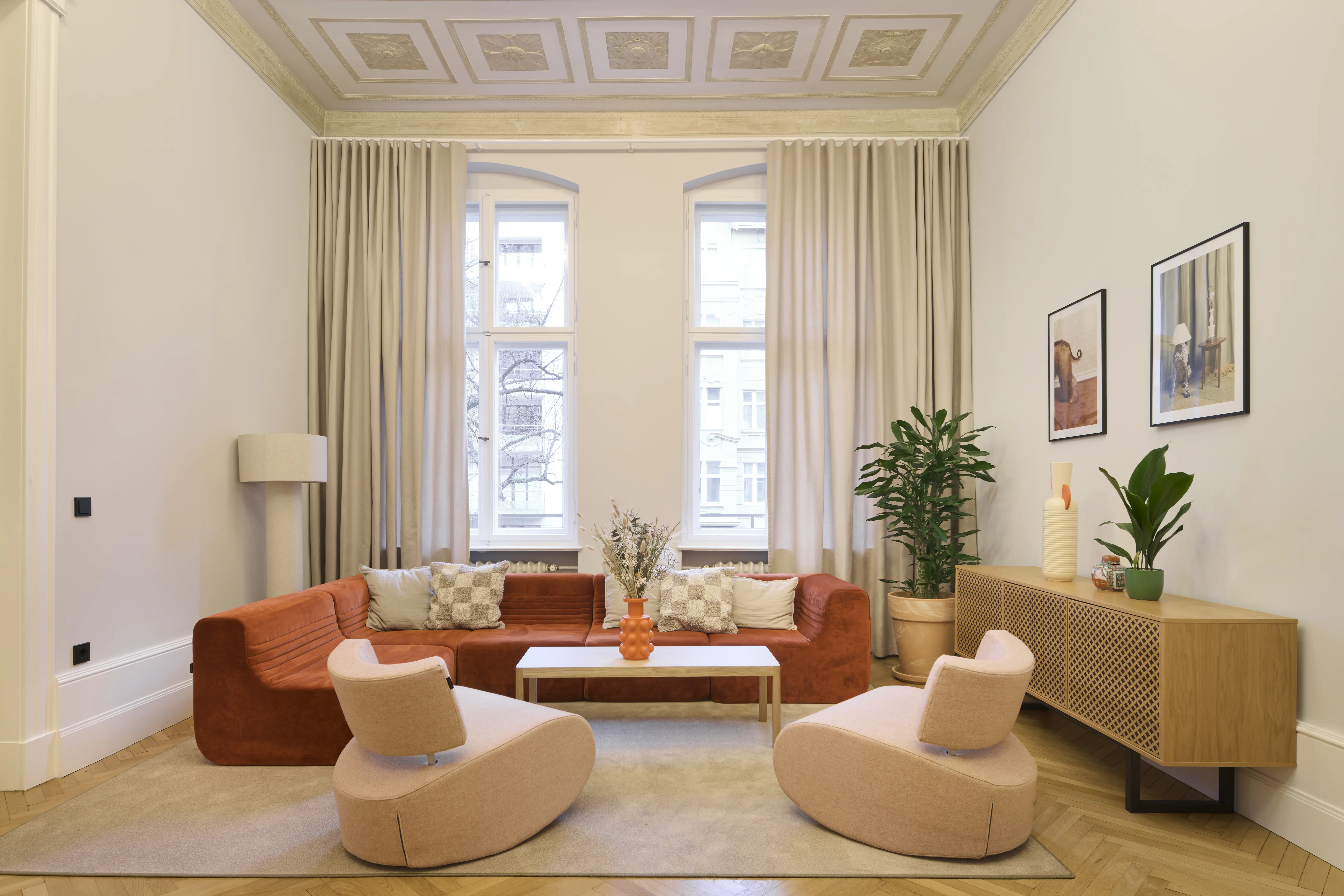Elegant lounge at INTERMATE BERLIN with a rust-red sofa, two blush-pink swivel armchairs, an oak sideboard, tall plants, framed artworks and high windows with beige curtains under a stucco ceiling.