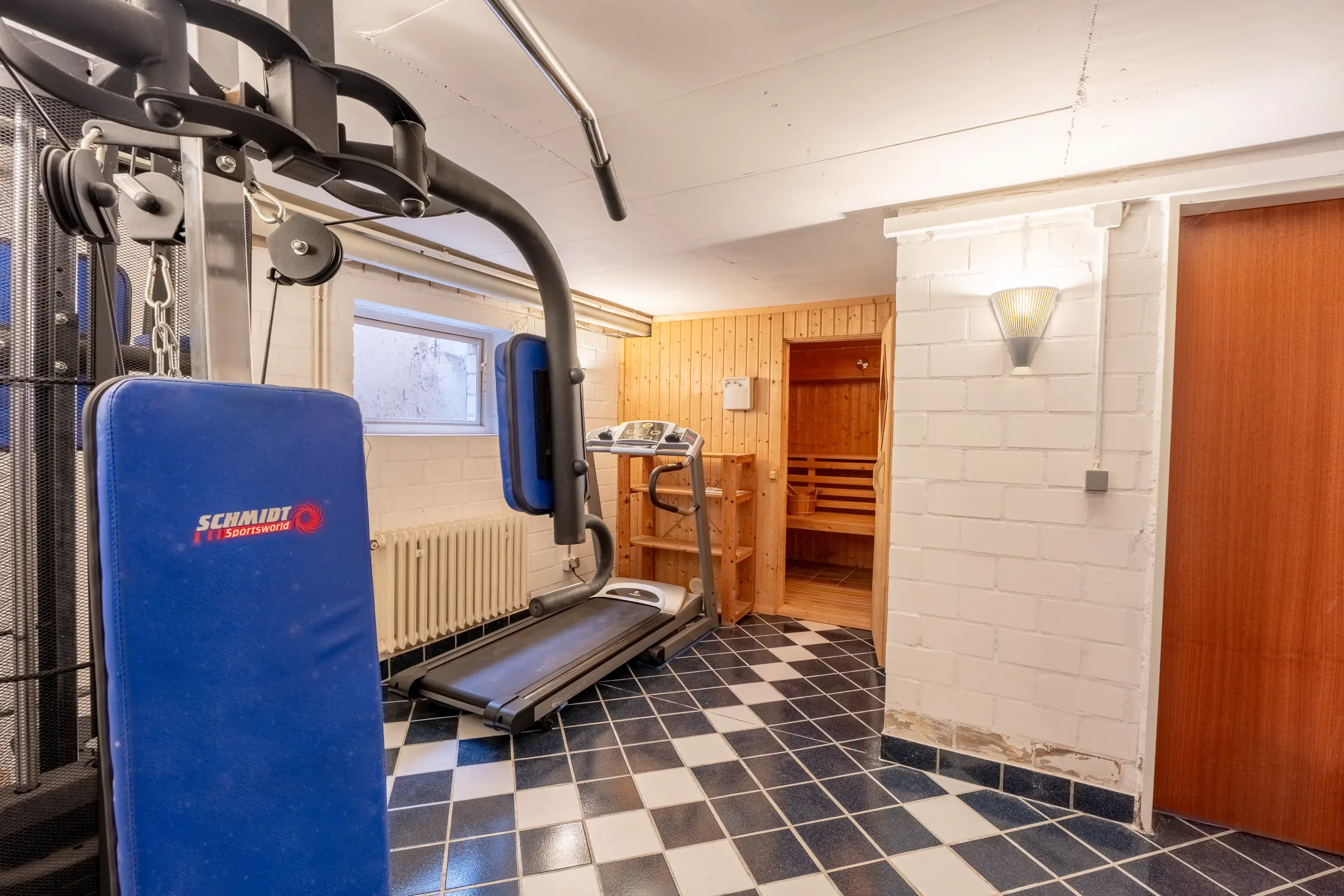 Gefliester Kellerraum mit Kraftstation, Laufband und Zugang zu einer holzverkleideten Sauna.