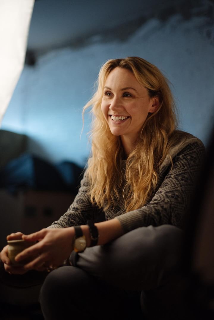 Close-up of a woman sitting on a couch and smiling warmly while sharing a testimonial about her singing lessons.