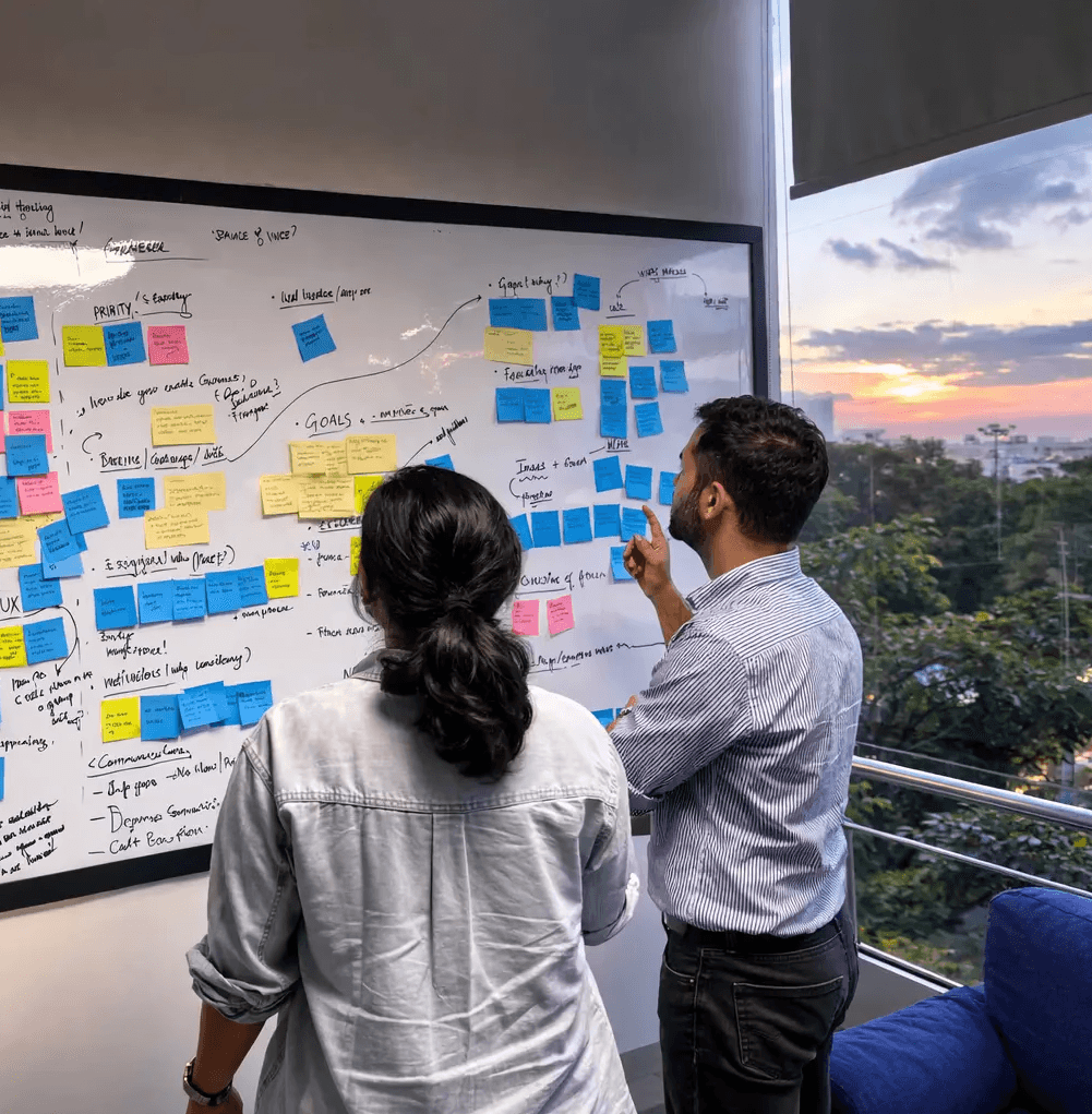 A man in a brown blazer and a woman in a light sweater sit at a desk with a laptop, discussing something. The room is dimly lit, creating a focused atmosphere.