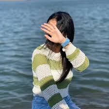 A person in a striped sweater is sitting by the water, partially shielding their face with their hand.