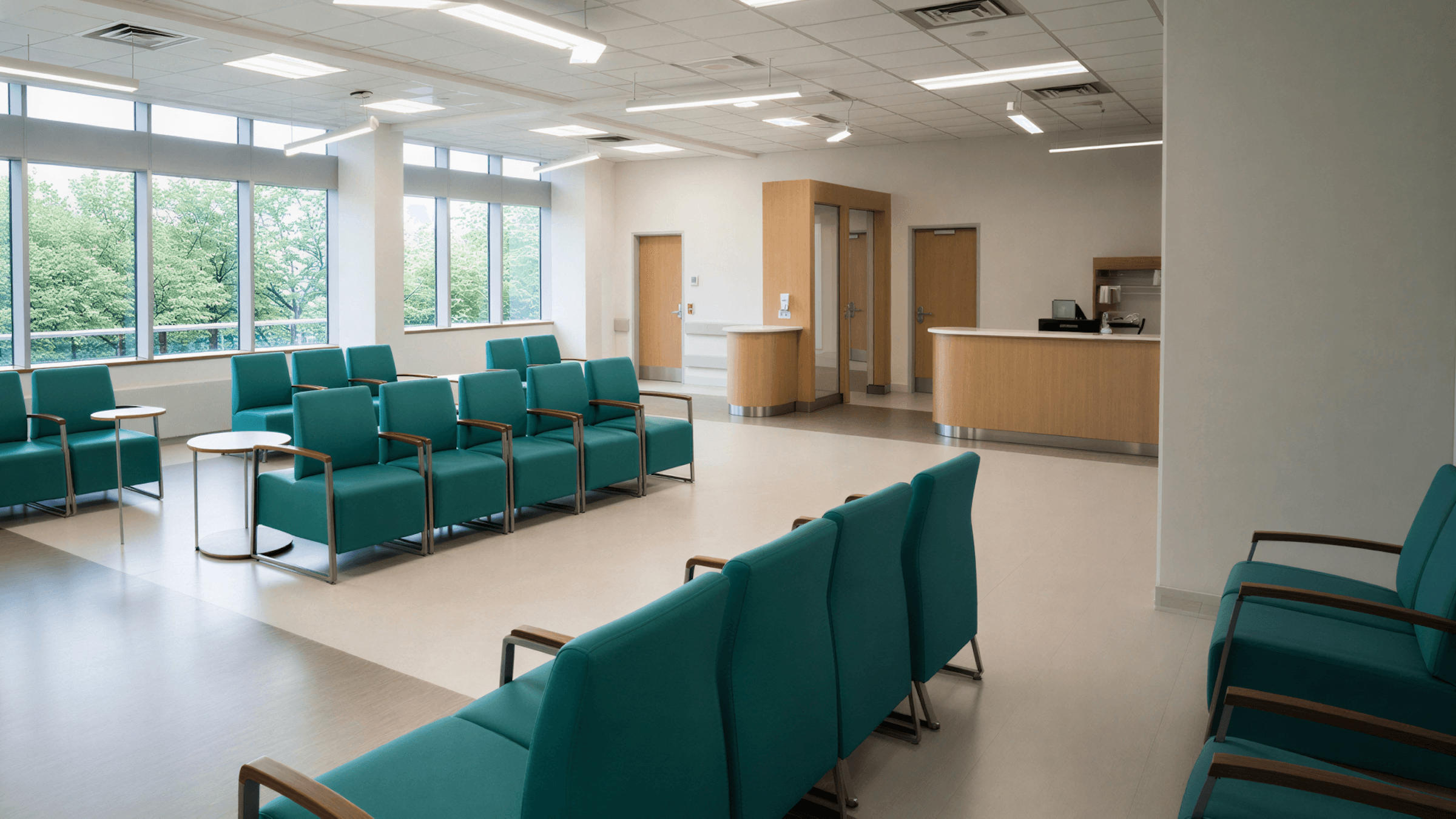 A modern waiting room features teal chairs, light walls, and large windows allowing natural light.