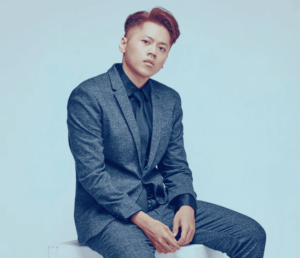 A man in a brown suit and cream silk scarf poses against a neutral background, exuding a composed and sophisticated demeanor.