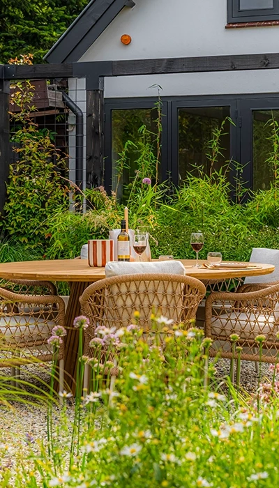 A cozy outdoor dining area surrounded by lush greenery and flowers, featuring a wooden table and stylish woven chairs.