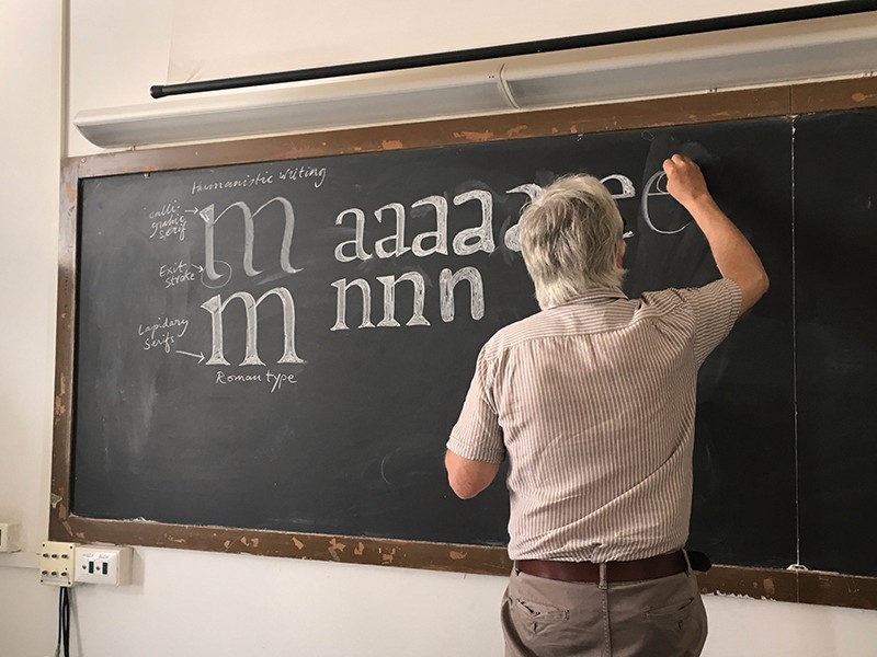 A man at a blackboard, teaching the typography and art of drawing letters.