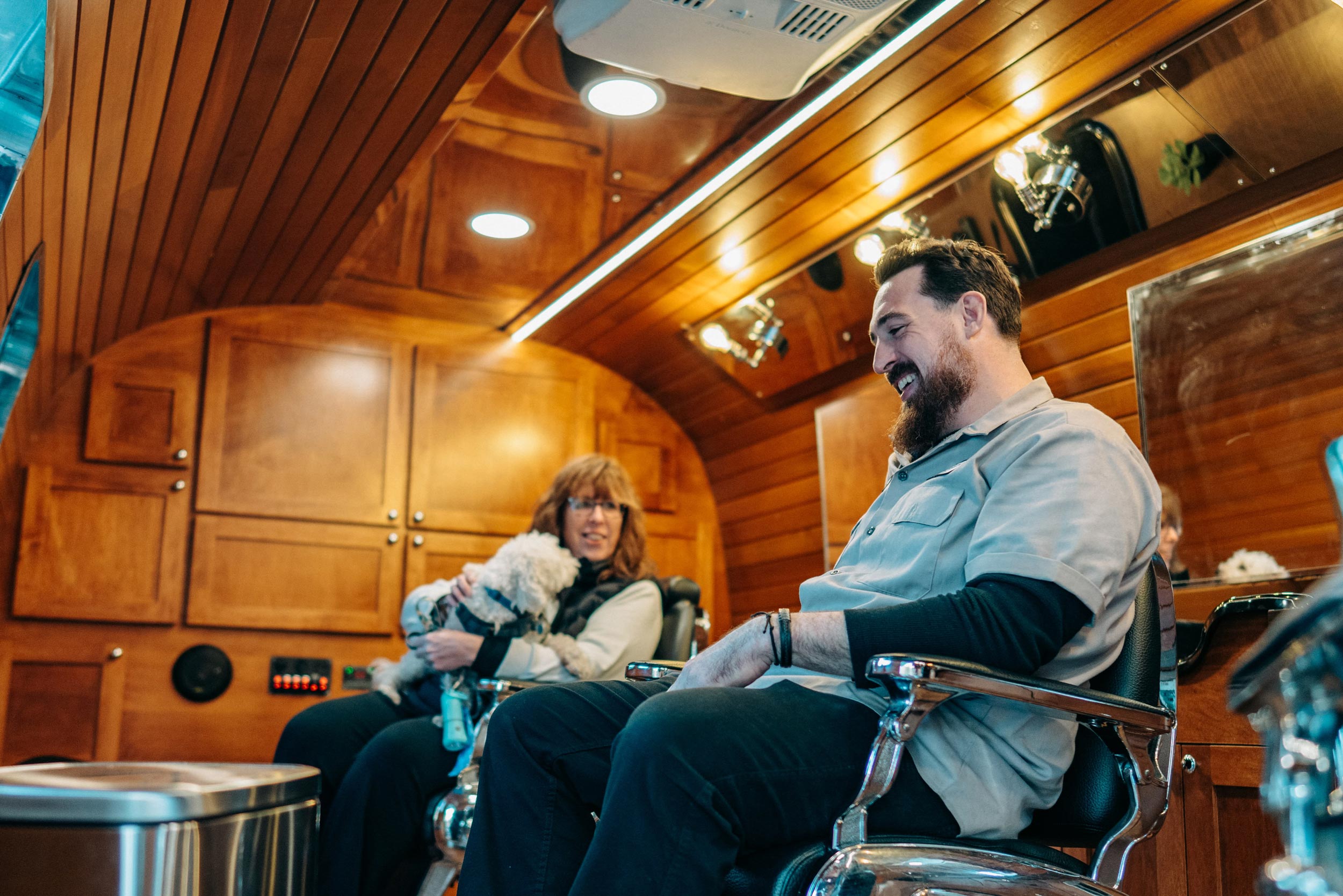 A man with a full beard, wearing a light-colored work shirt over a dark long-sleeved shirt, is seated in a chrome barber chair and laughing. To the left, another person is seated on a bench, holding a small white, fluffy dog. The interior of the space is finished with warm, rich wood paneling on the walls and ceiling, with modern strip lighting.