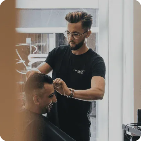 A barber wearing a black t-shirt and glasses skillfully cuts the hair of a seated customer in a modern, well-lit barbershop.