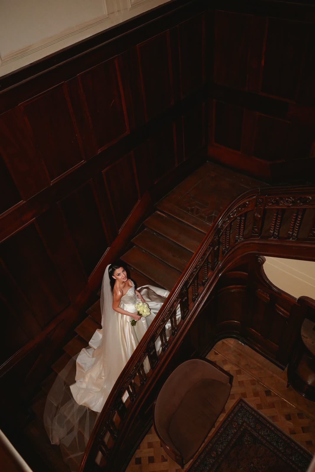 Bride in a white gown on a dark wooden staircase seen from above