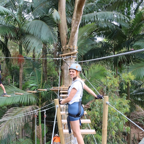 شخص يرتدي خوذة ومعدات أمان يقف على جسر حبال في أعالي الأشجار في غابة استوائية خصبة.