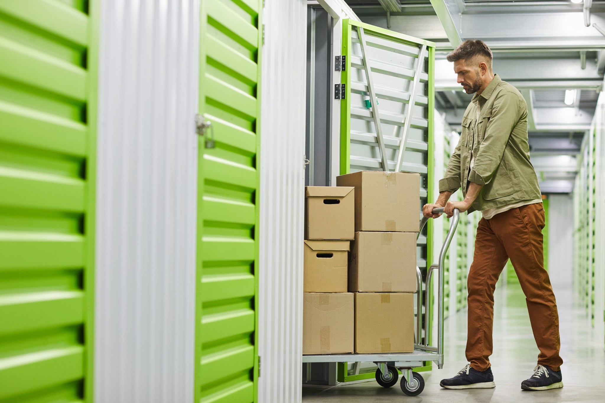 Man with dolly pushing boxes in a self-storage facility with bright green doors.