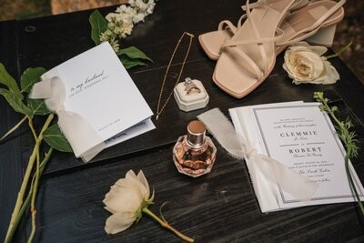 Wedding stationery, pale sandals, rings, perfume and white flowers arranged on a dark wooden surface.