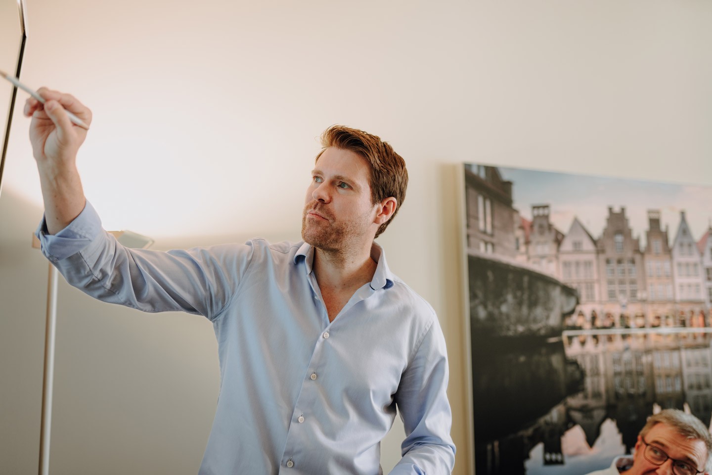 Männlicher Geschäftsführer im hellen Hemd präsentiert Inhalte an einem Whiteboard vor einem Foto im Hintergrund.