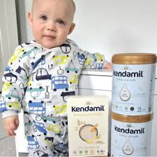 A smiling baby in pajamas stands next to cans of baby formula, holding one of the boxes with a happy expression.