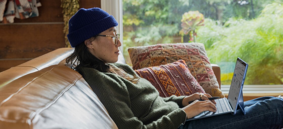 Woman in blue hat and sweater sitting on couch with laptop.