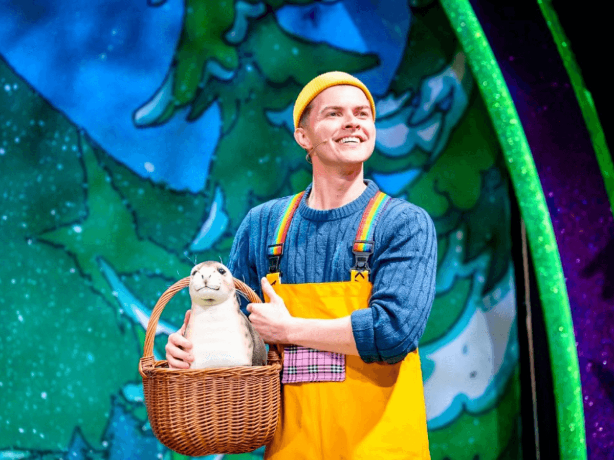 ビューティ・アンド・ザ・ビーストで黄色のオーバーオールを着た笑顔の男性が舞台で籠を持つシーン『ホーニー・ラブ・ストーリー』。