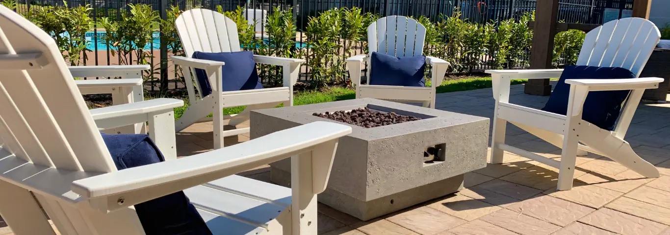 Four white Adirondack chairs with blue cushions arranged around a square stone fire pit.