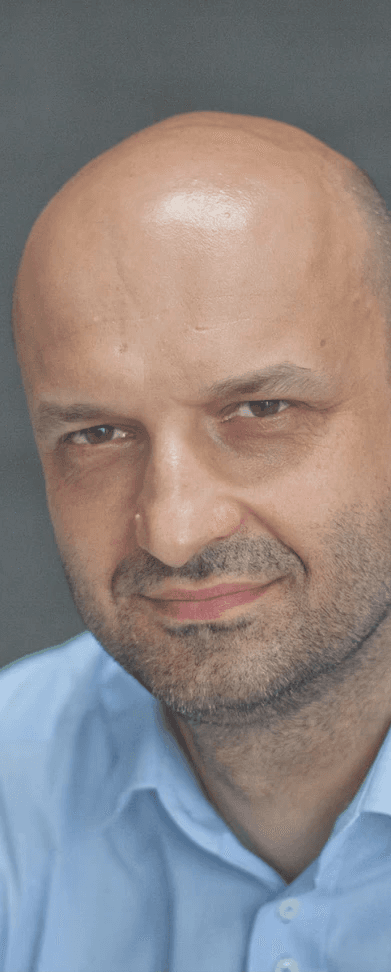 Zdjęcie przedstawia kobietę o jasnych, falowanych włosach i okularach w ciemnych oprawkach. Ma na sobie czarną bluzkę z wzorem i patrzy prosto w obiektyw. Tło jest jednolite, ciemne.