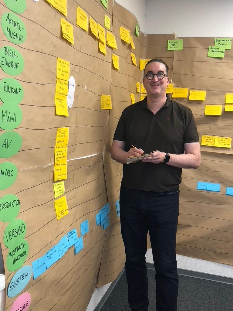 Bernd Winkhaus steht vor einer mit bunten Haftnotizen bedeckten Wand, lächelt und hält ein Gerät in der Hand.