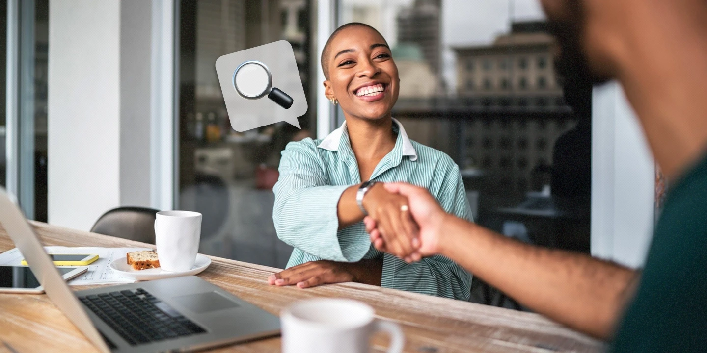 Mulher sorridente aperta a mão de outra pessoa em um ambiente de trabalho, com um laptop sobre a mesa. Um ícone de lupa flutua ao lado dela simbolizando o monitoramento do WhatsApp no ambiente corporativo