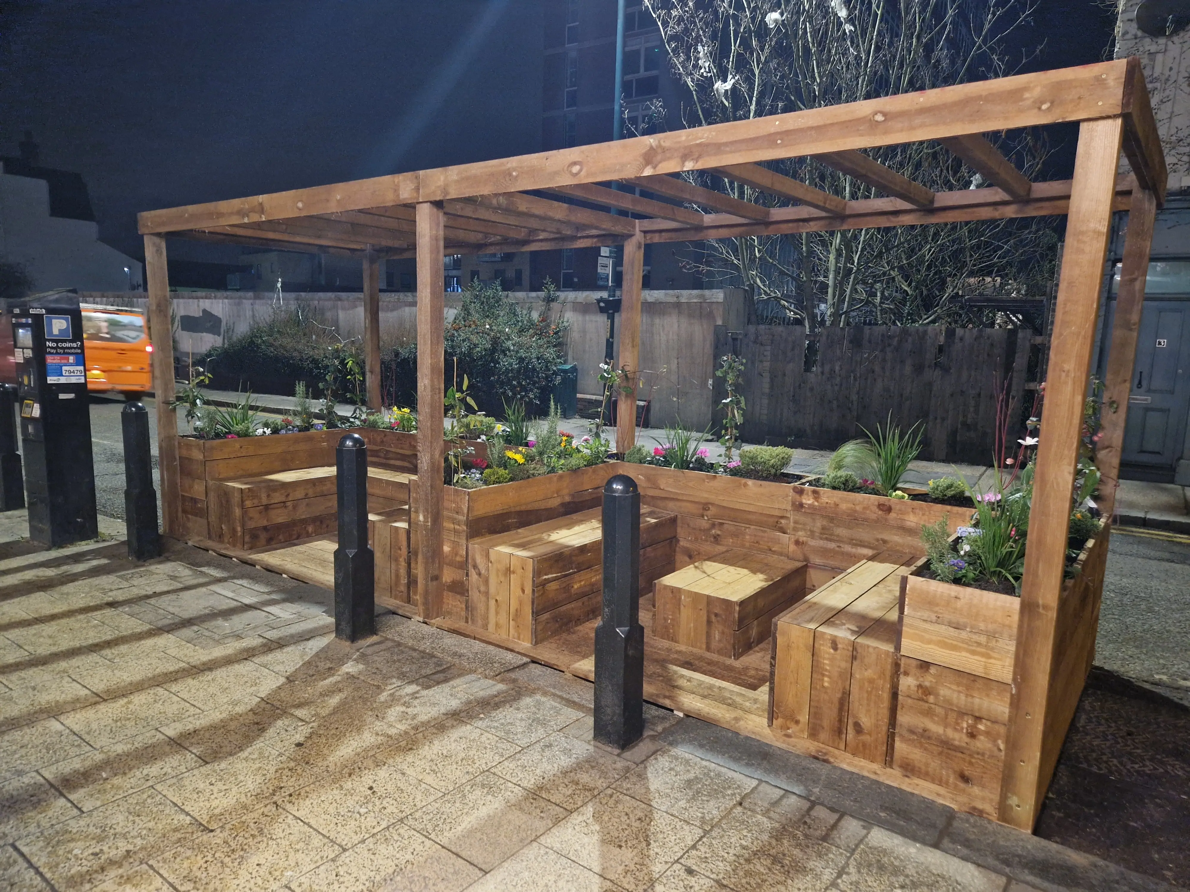 A wooden outdoor seating area with benches surrounded by plants, illuminated at night.