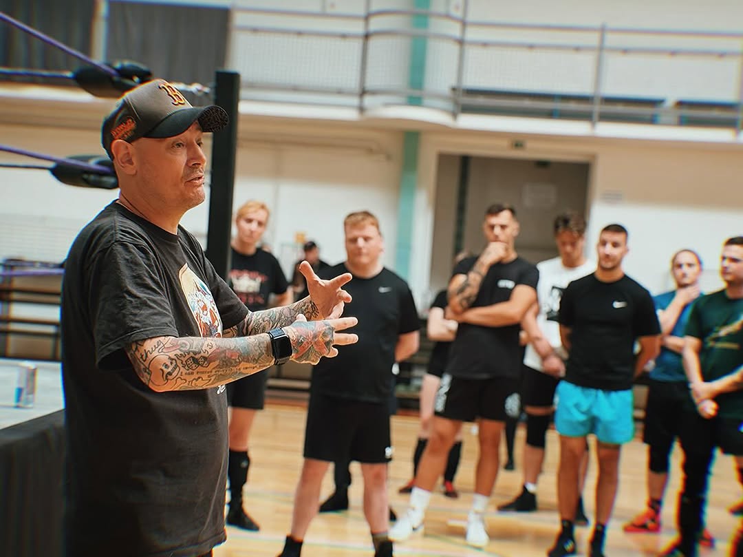A group of people wearing aprons gather around a wooden table, shaping and scoring round loaves of bread dough covered with flour, with bowls and a sunlit window in the background.