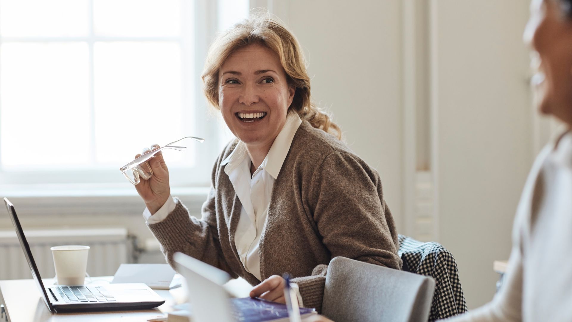 A teacher pauses at her desk, laptop open, glasses in hand