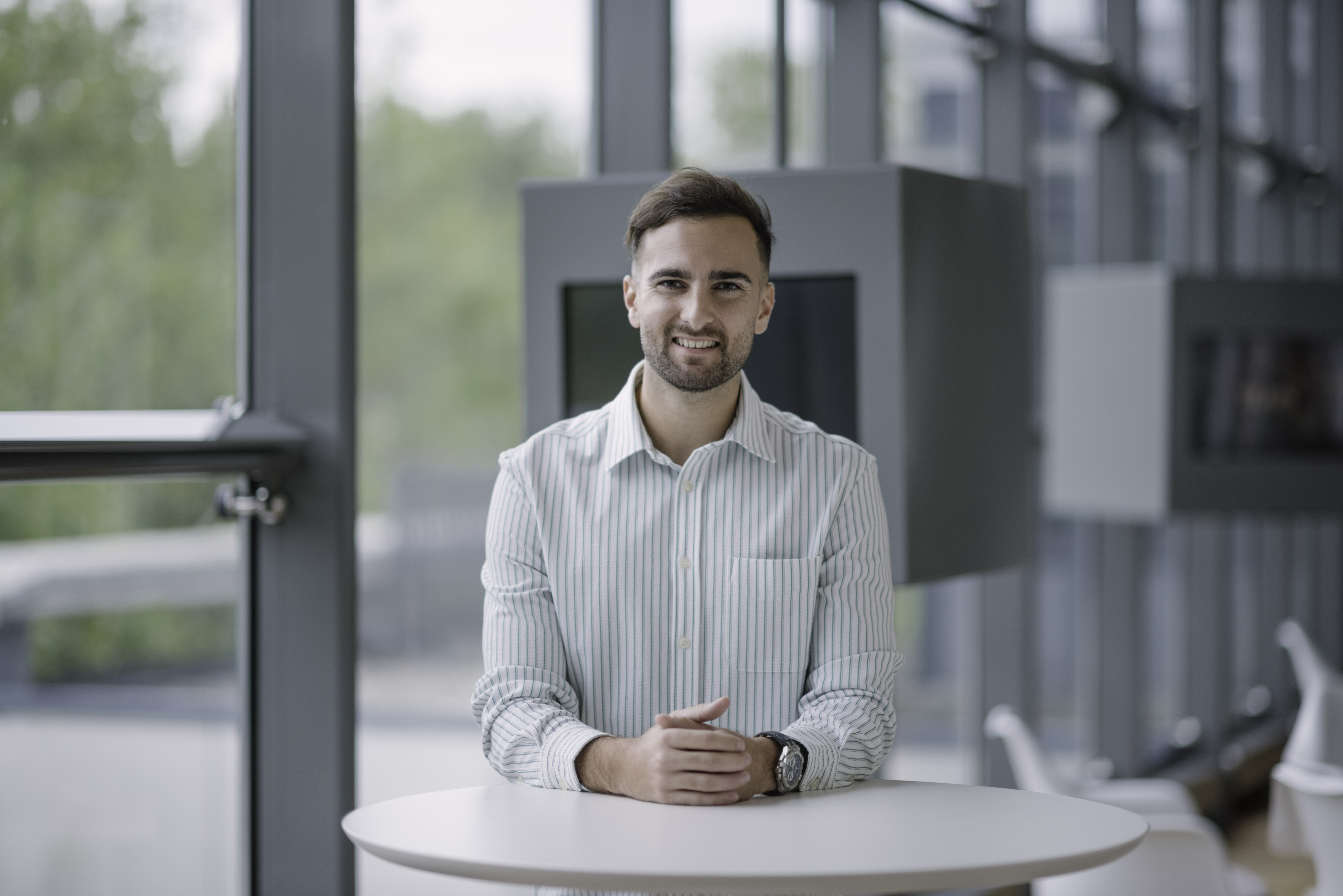 Junger Mann aus dem Team steht selbstbewusst an einem Tisch in einem hellen, modernen Büro.