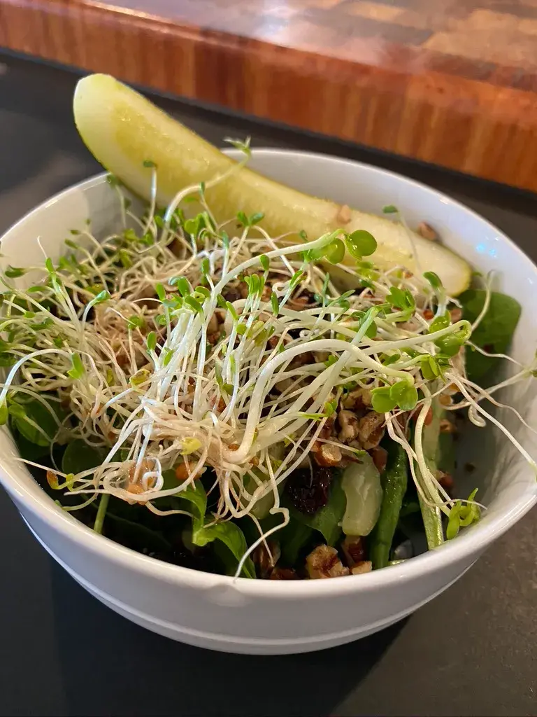 A fresh green salad topped with crunchy sprouts, chopped nuts, and accompanied by a crisp dill pickle spear in a white bowl on a dark surface.