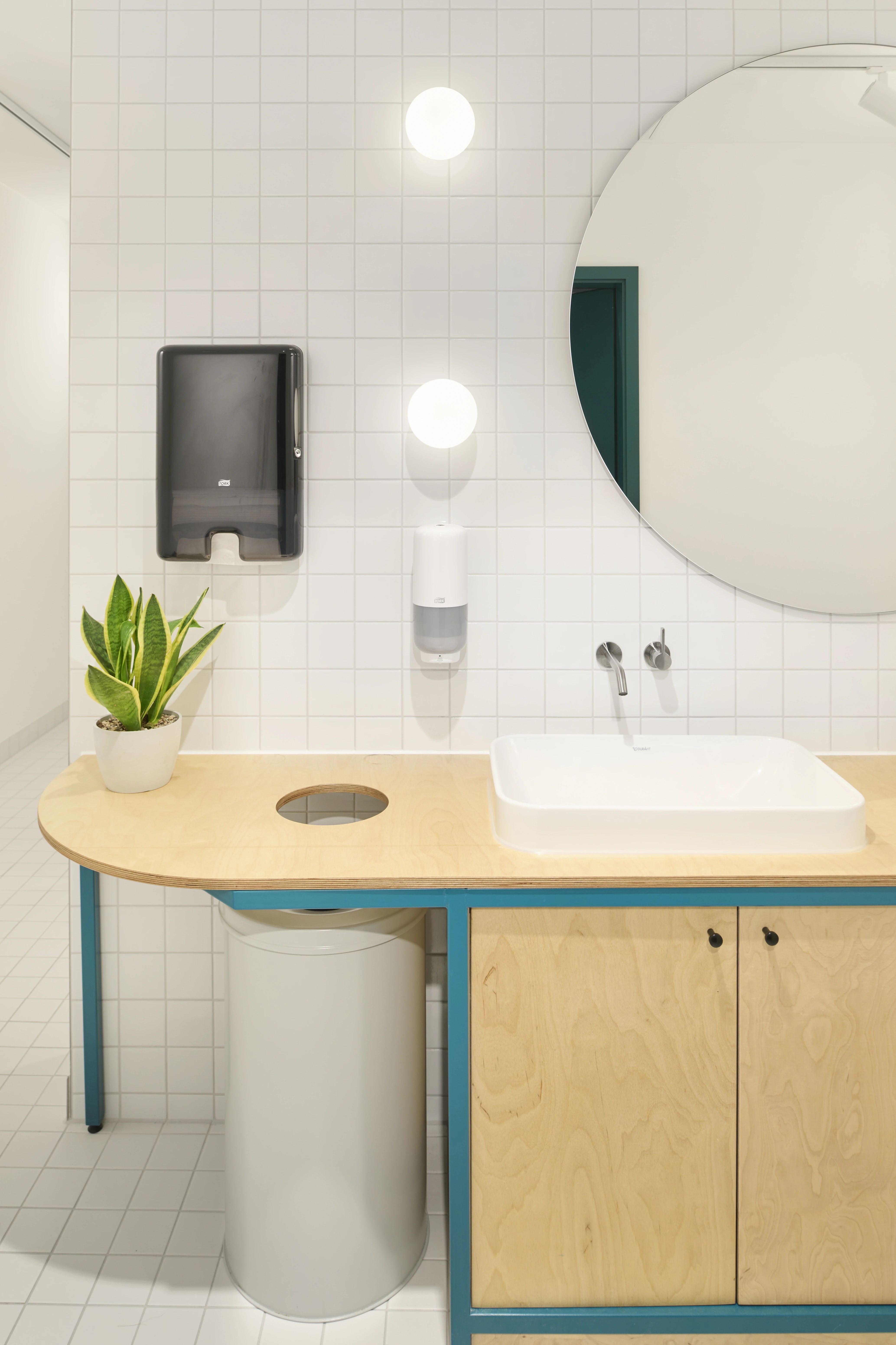 Washroom at HELLOFRESH GYM with a white basin on a rounded plywood counter with teal frame, round mirror, white tiles and globe wall lights.