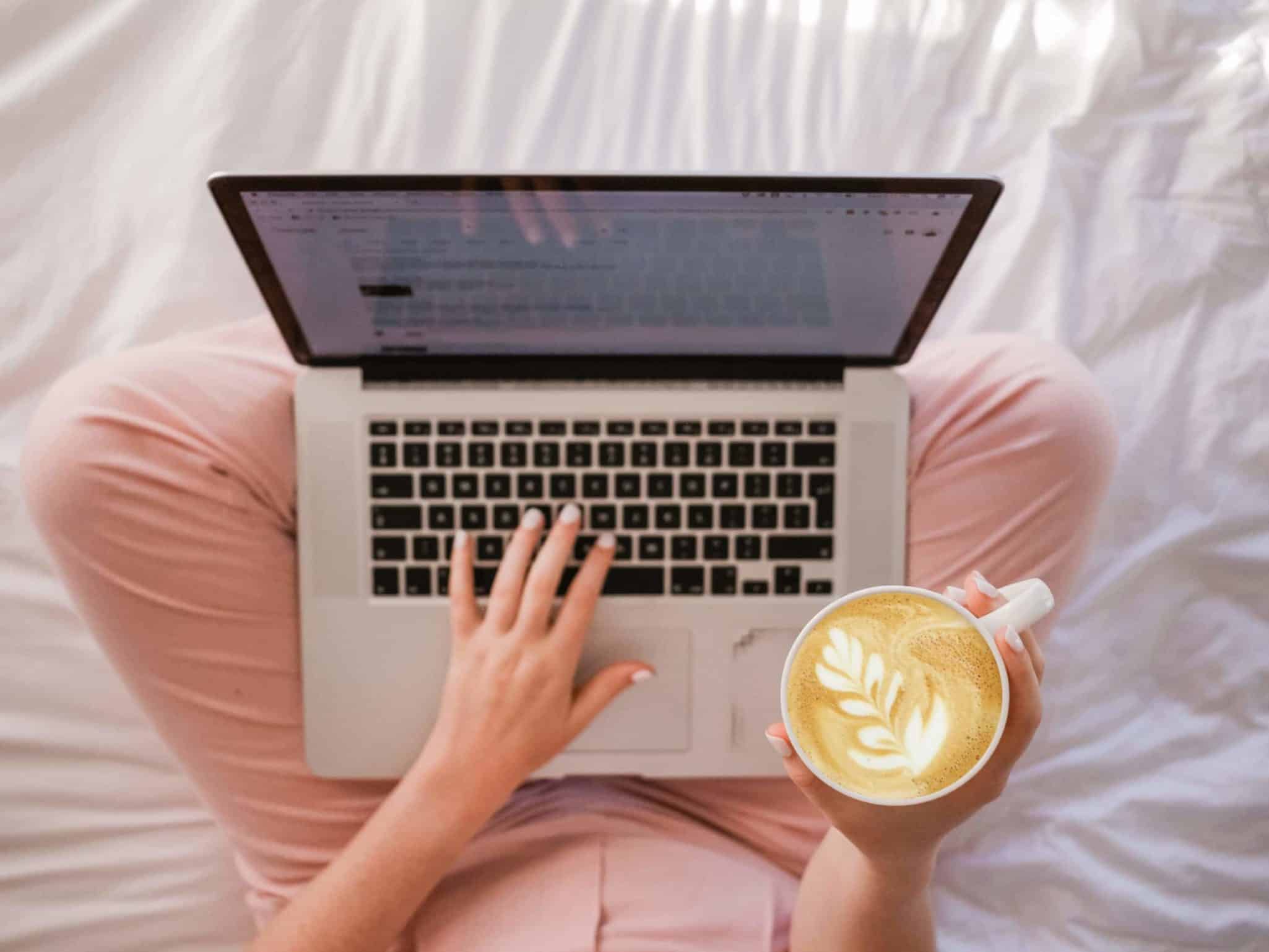 Person using laptop on bed with latte art coffee in hand.