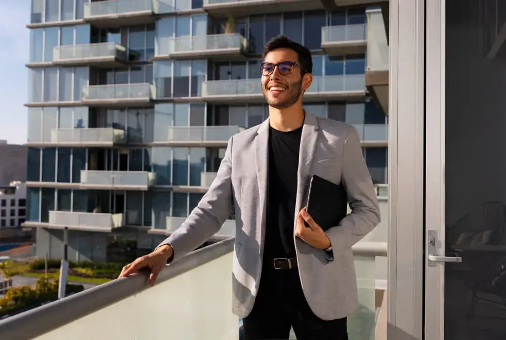 A confident real estate investor stands on a modern apartment balcony, smiling and holding a tablet with city buildings in the background. The image represents success, growth, and financial confidence made possible through Investment Loans from Chris Lewis Home Loans.