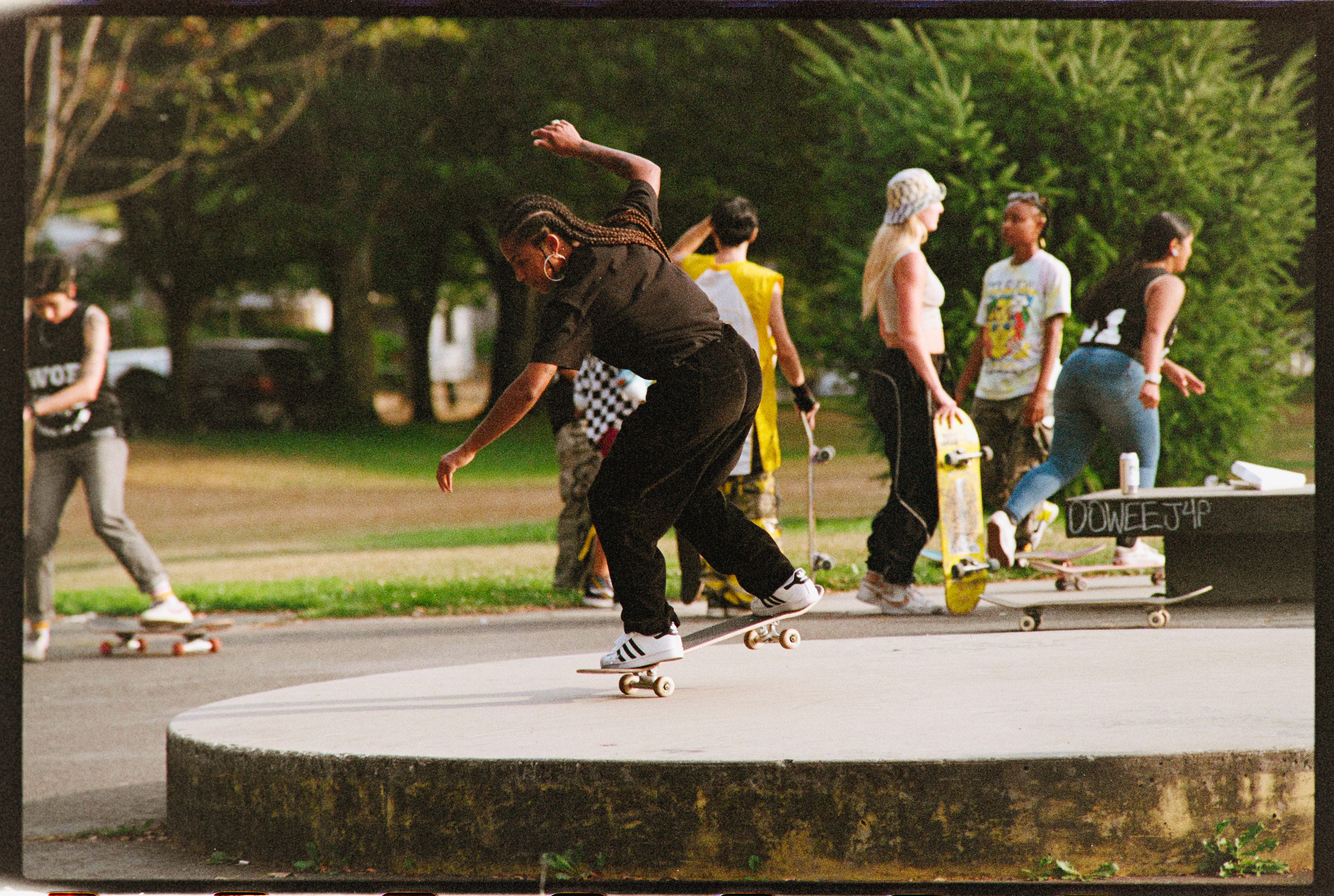 A skatepark scene with diverse skaters practicing tricks. A person in the foreground skates on a platform, while others socialize in the background. Energetic and vibrant atmosphere.