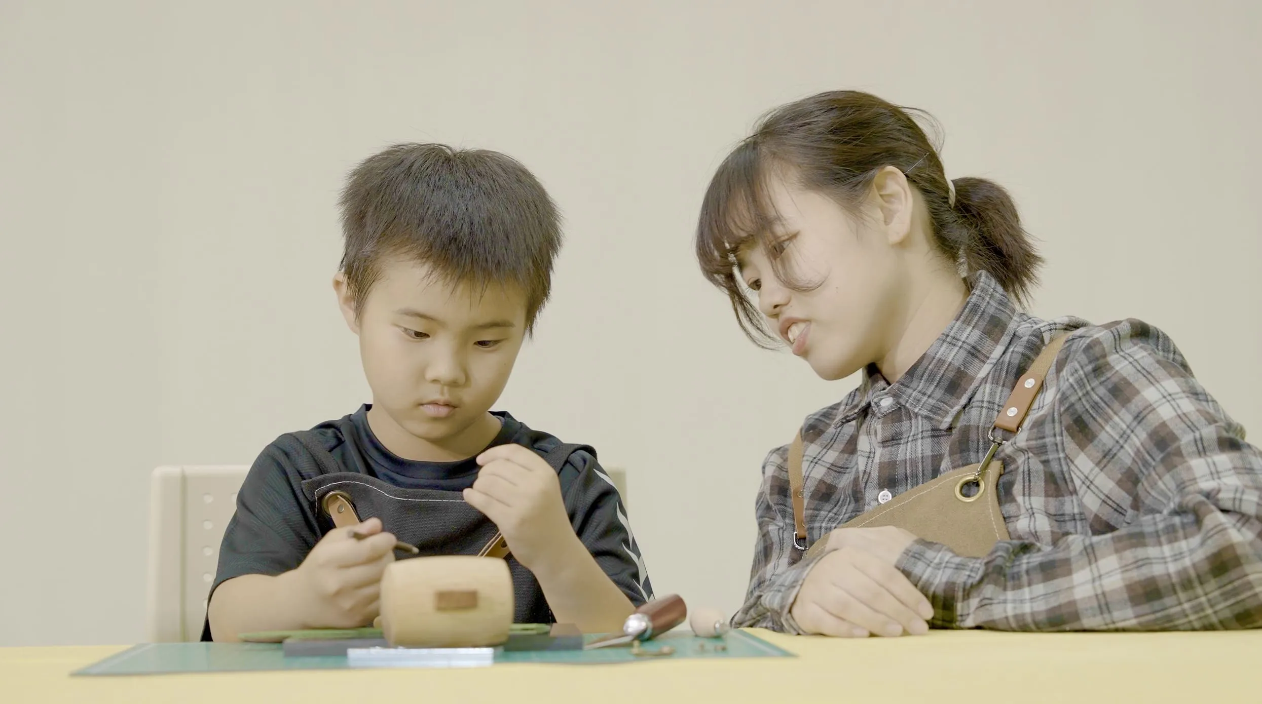 An instructor at our kids academy provides focused, one-on-one guidance to a young student during a hands-on craft workshop. The boy concentrates intently on his project at a yellow table while the teacher leans in to assist.